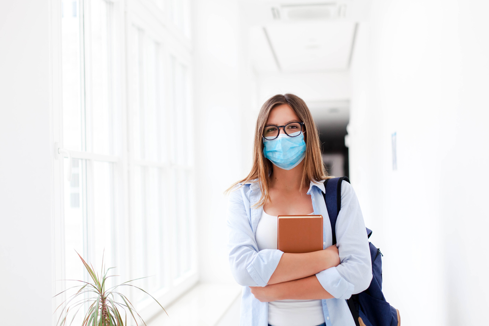 Student in protective face mask in empty college indoors. Young woman going to exams in high school. Girl with backpack and book in university corridor. Social distancing during quarantine.