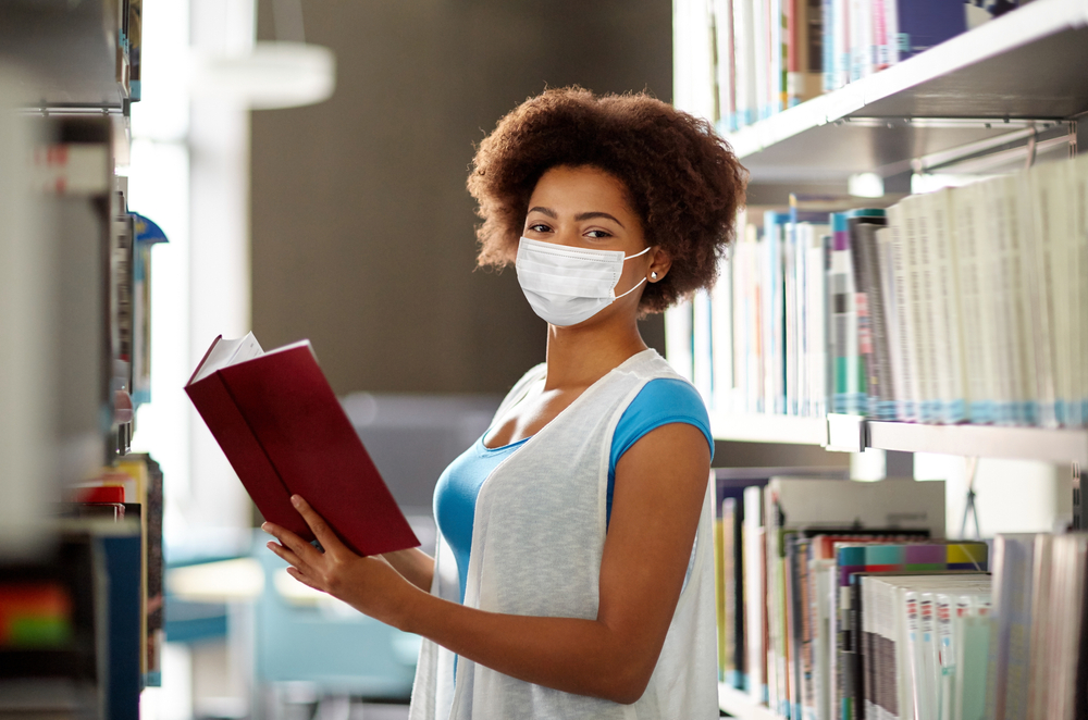 african american student girl wearing face protective medical mask for protection from virus disease reading book at library